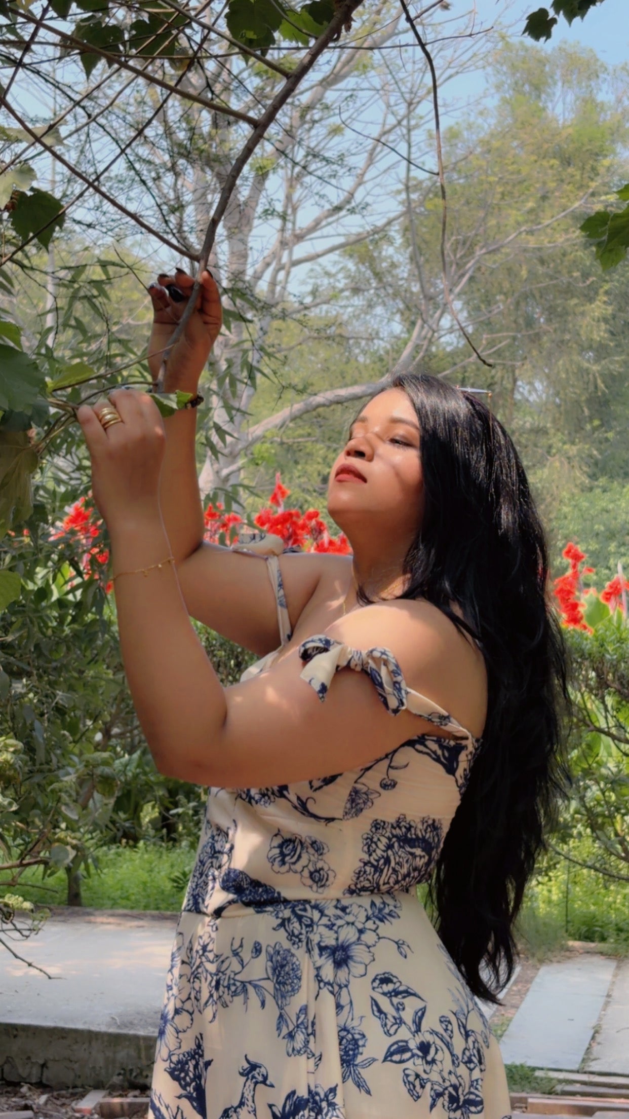 Woman in a floral dress standing outdoors with greenery and flowers in the background