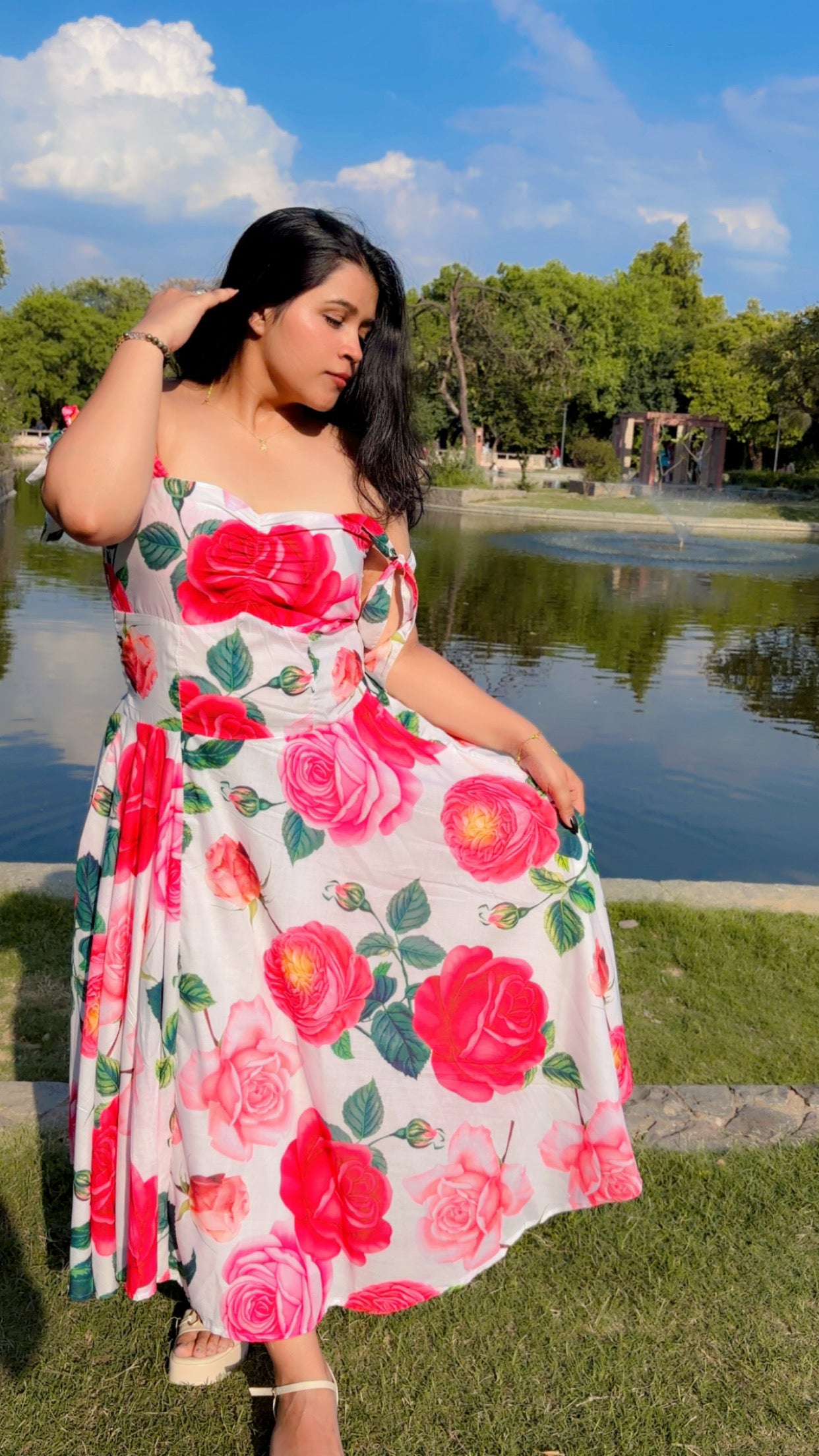 Woman in a floral dress sitting by a pond with trees and blue sky in the background