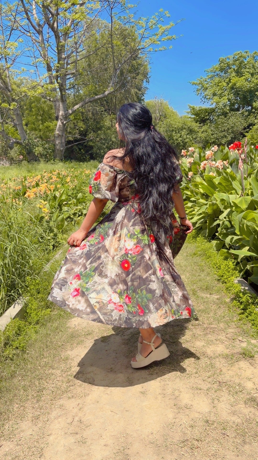 Woman in a floral dress walking through a garden with flowers and trees.